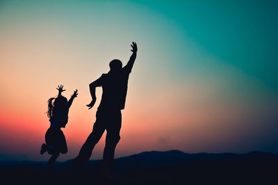 Silhouette man and girl jumping with arms raised against clear sky during sunset