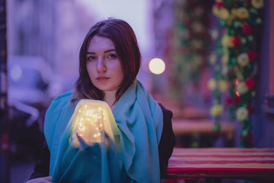 Portrait beautiful woman with blue scarf and glowing jar in city at dusk