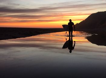 Silhouette man carrying surfboard while walking on beach during sunset