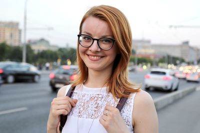 Portrait of smiling young woman wearing glasses and holding school bag in city jam