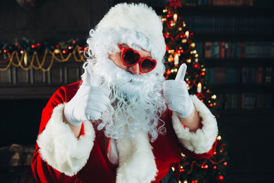 Portrait of man wearing santa claus costume while gesturing thumbs up at home