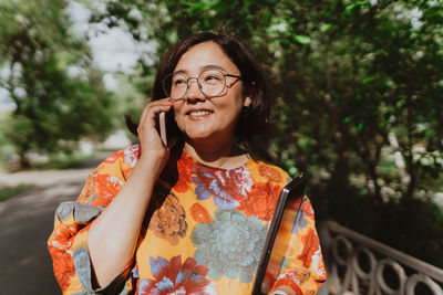 Cheerful young woman with glasses talking on a mobile phone in lush green park.