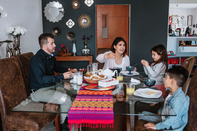 Positive young latin american mother and father with children having delicious breakfast and communicating during weekend at home