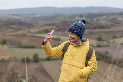 Happy little hiker having fun while taking a selfie with smart phone during autumn day on a hill.