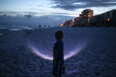 Boy holding flashlight while standing at beach against sky