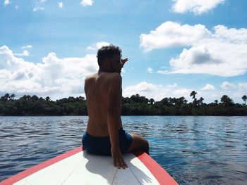 Rear view of shirtless man sitting on lake against sky