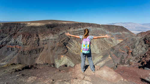 Rear view of woman with arms outstretched standing on rocky mountain against clear sky