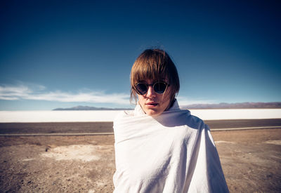 Portrait of woman wearing sunglasses wrapped in blanket standing on land against sky during sunny day