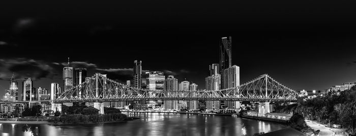 Illuminated bridge over river by buildings against sky at night