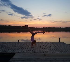 Full length of girl practicing handstand on pier over lake against sky during sunset