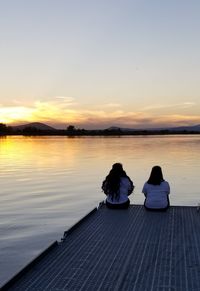 Rear view of women sitting on pier at lake against sky during sunset