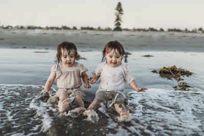 Front view of toddler sisters sitting and splashing in water at beach