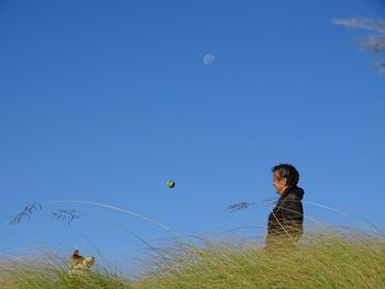 Side view of woman throwing ball towards dog on grassy field against clear blue sky