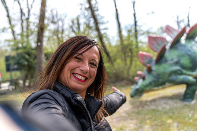 Smiling middle aged woman taking a selfie in front of dinosaur models in a jurassic park