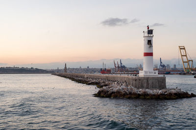 Flock of birds perching on groyne by lighthouse against sky during sunset