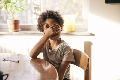 Multiracial boy covering eyes with hands while sitting on chair at home