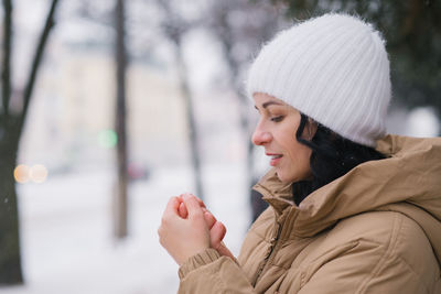 A young girl in a white hat warms her hands. it stands in the city in winter under the snow