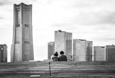 Rear view of couple sitting by retaining wall against cityscape