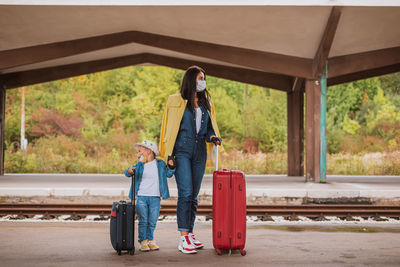 Full length of mother with son wearing mask standing on railroad station platform