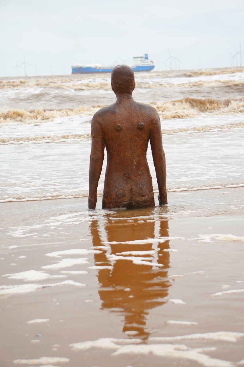 REAR VIEW OF SHIRTLESS MAN STANDING ON WET SHORE