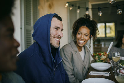 Smiling mature man wearing hood sitting with female friends at dining table in backyard patio