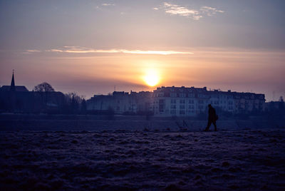 Silhouette man walking on field against sky during sunset in city