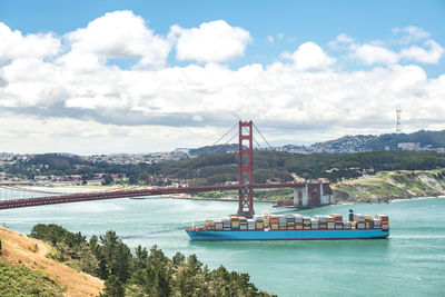 Container ship in river crossing golden gate bridge against cloudy sky