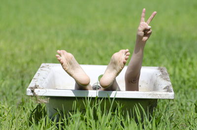 Low section of woman in bathtub showing victory sign on grassy field