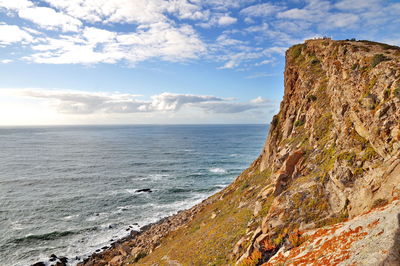 Scenic view of sea by rock formation against cloudy sky during sunny day