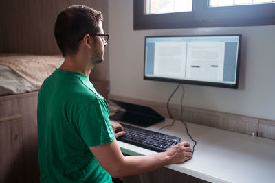 Side view of male blogger in eyeglasses editing text on monitor while typing on keyboard in house room