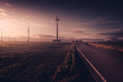 Road by field against sky during sunset with wind turbines