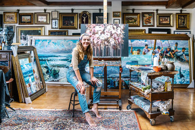 Cheerful female artist in denim overall looking at camera while sitting near easel and painting in creative atelier