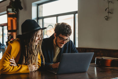 Smart man in glasses and stylish woman in hat looking at monitor while man typing on laptop in cafe