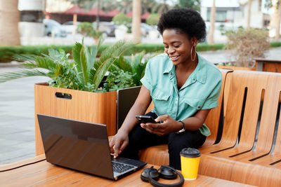 Smiling woman using mobile phone and laptop while sitting at outdoor cafe