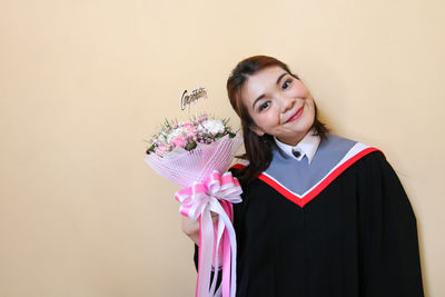 Portrait of young woman wearing graduation gown holding bouquet against wall