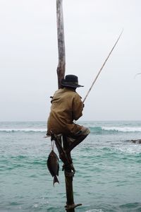 Rear view of man fishing in sea while sitting on bamboo against sky