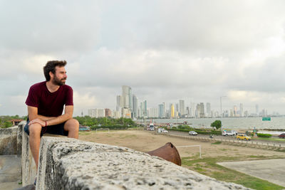 Side view of woman sitting on retaining wall against sky