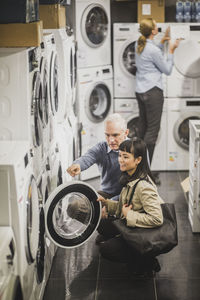 High angle view of mature owner pointing to washing machine while female customer crouching in electronics store