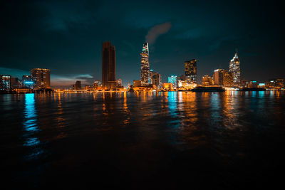 Illuminated city by river against sky at night