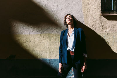 Thoughtful female wearing elegant clothes standing sunlight and casting shadow on shabby wall and looking away