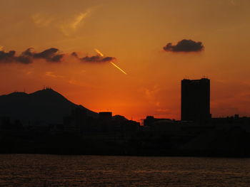 Silhouette cityscape by sea against sky during sunset