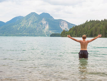 Teenager boy stands in water and salutes with raised arms. swimming in alpine lake, herzogstand peak