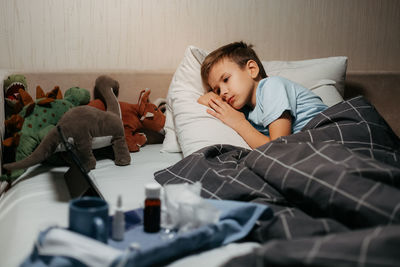 Very sad ill child laying in bed with toys and tablet. some medicines are nearby