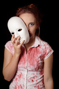 Close-up portrait of young woman holding mask over black background