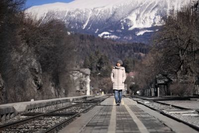 Full length of woman wearing warm clothing while walking on railroad station platform against mountains during winter