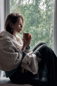 Side view of woman having drink while sitting on window