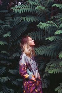 Side view of young woman with hands in pockets standing by trees