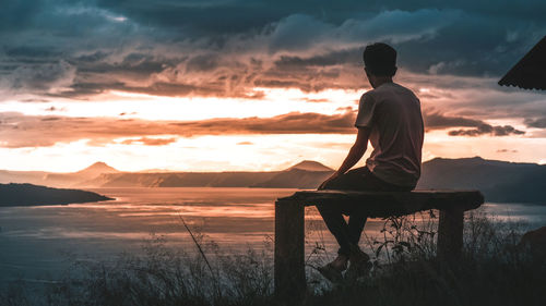 Rear view of man sitting on shore against sky during sunset