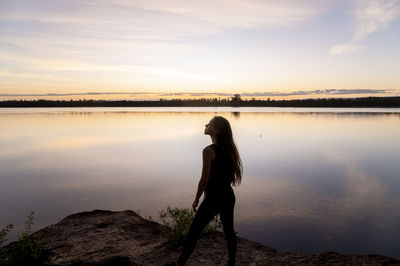 Rear view of man standing by lake against sky during sunset