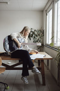 Full length of mature woman sitting on table while practicing guitar in classroom